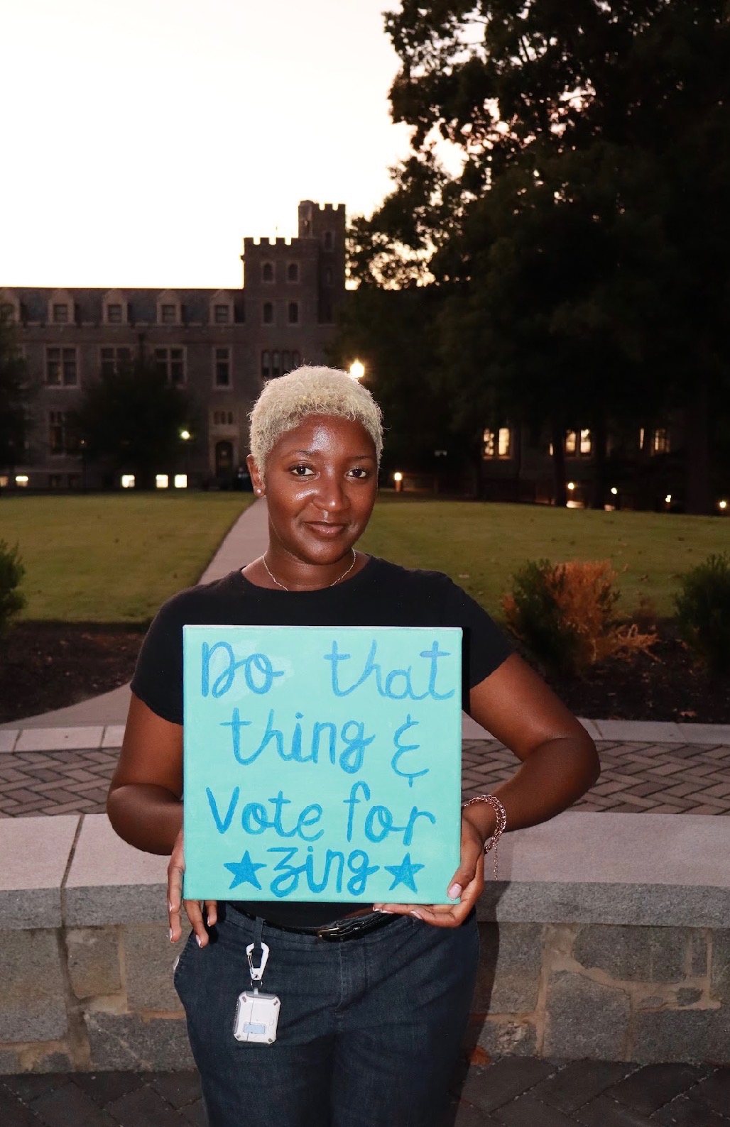 Nzinga on campus at dusk holding a painted sign reading Do that thing and Vote for Zing