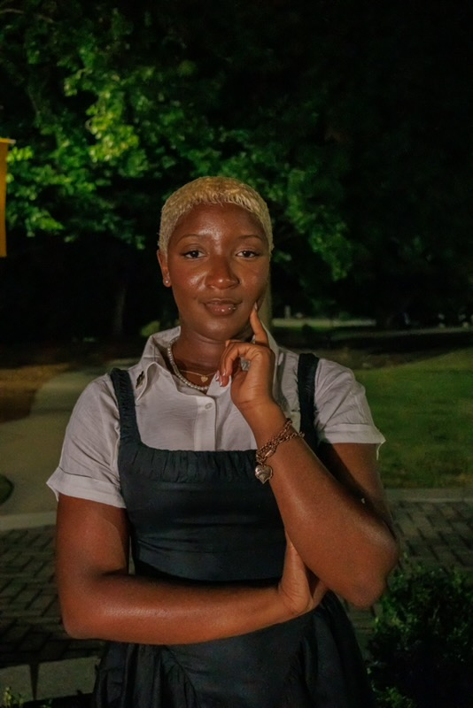 Nzinga in a white shirt and black dress, resting her chin thoughtfully on her hand outdoors at night