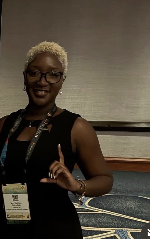 Nzinga wearing glasses and a conference lanyard, raising one finger