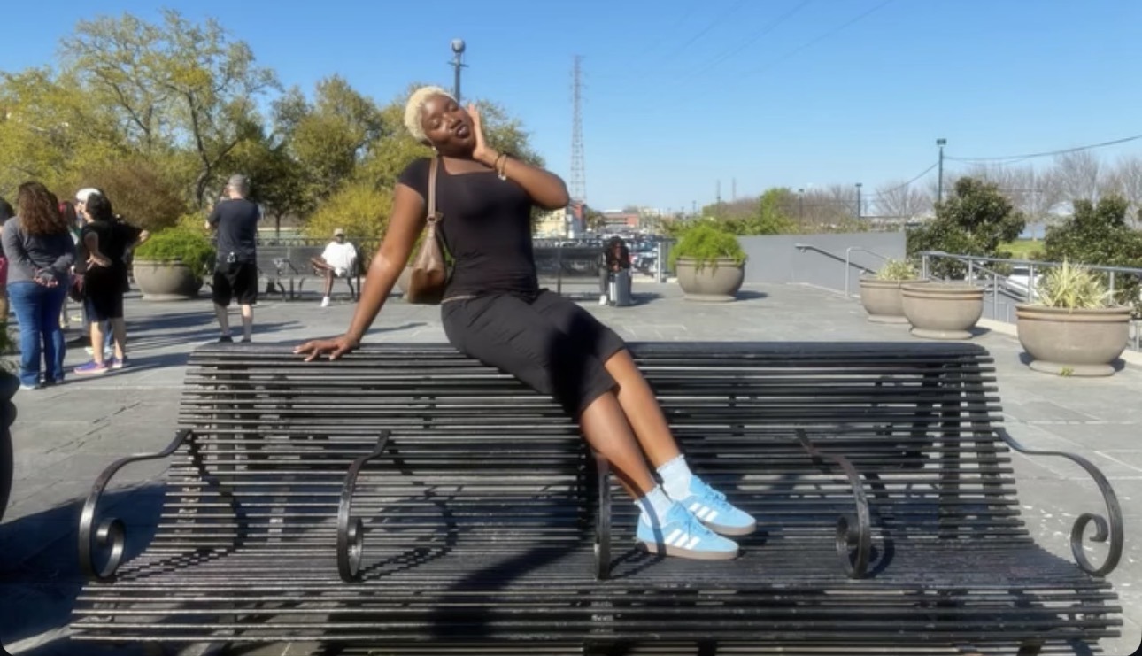 Nzinga sitting on an outdoor bench in black clothing and blue sneakers